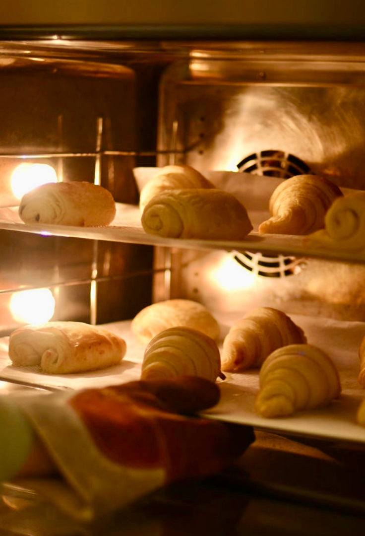 Croissants, pains au chocolat, and pains aux raisins rising and turning golden on multiple racks inside a professional convection oven with warm light during a croissant baking class at Maison Fleuret Paris