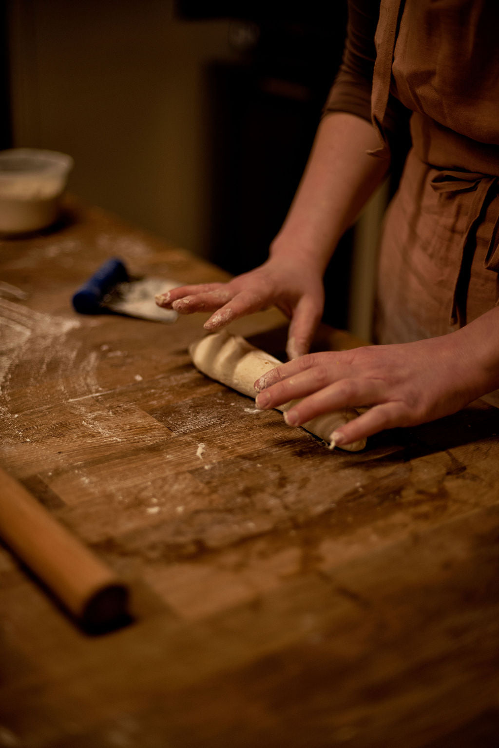 Participant in apron shaping a scored baguette dough by hand on a flour-dusted wooden table with a bench scraper and rolling pin during a bread class at Maison Fleuret Paris