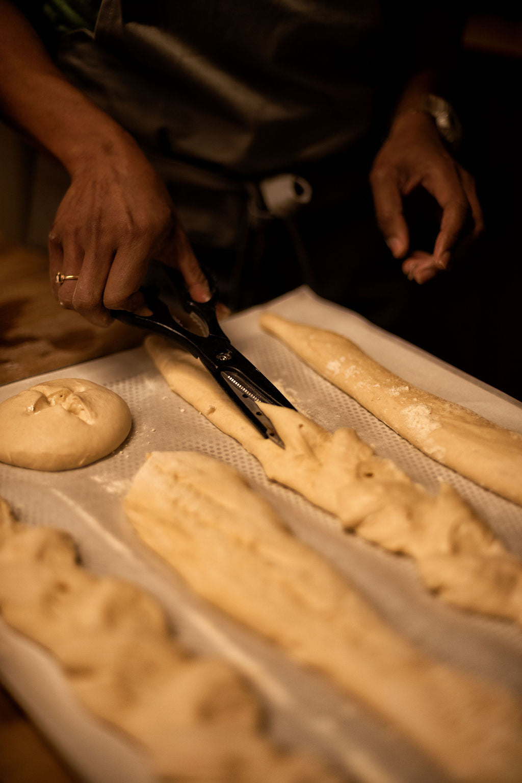 Baker snipping bread dough with scissors to create an epi de ble wheat stalk shape alongside baguettes and rolls on a baking tray during a bread class at Maison Fleuret Paris