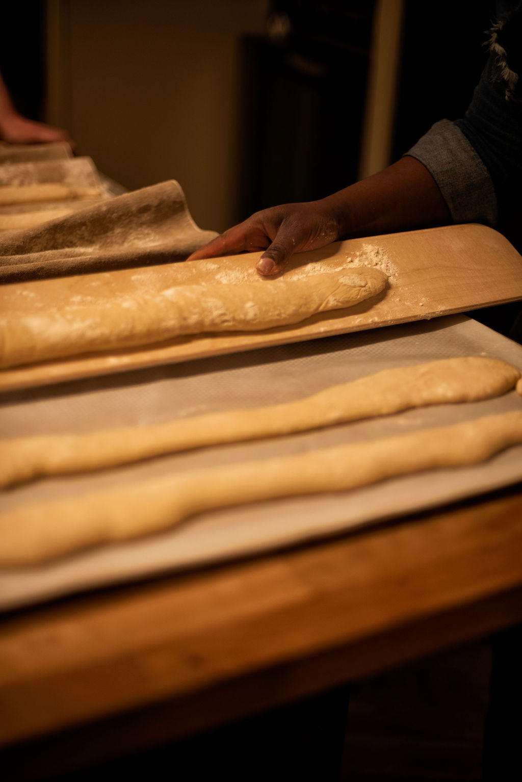 Participant transferring a shaped baguette dough onto a wooden peel with flour-dusted hands, with rows of formed baguettes on parchment below during a bread baking class at Maison Fleuret Paris