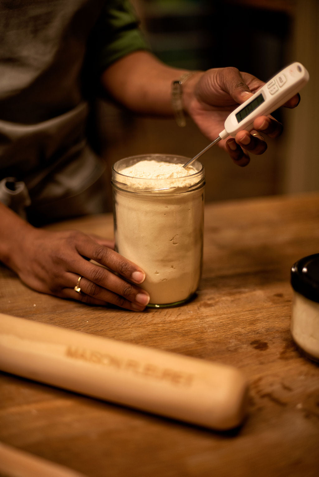 Baker checking the temperature of a sourdough starter in a glass jar with a digital thermometer next to a branded Maison Fleuret rolling pin during a bread baking class in Paris