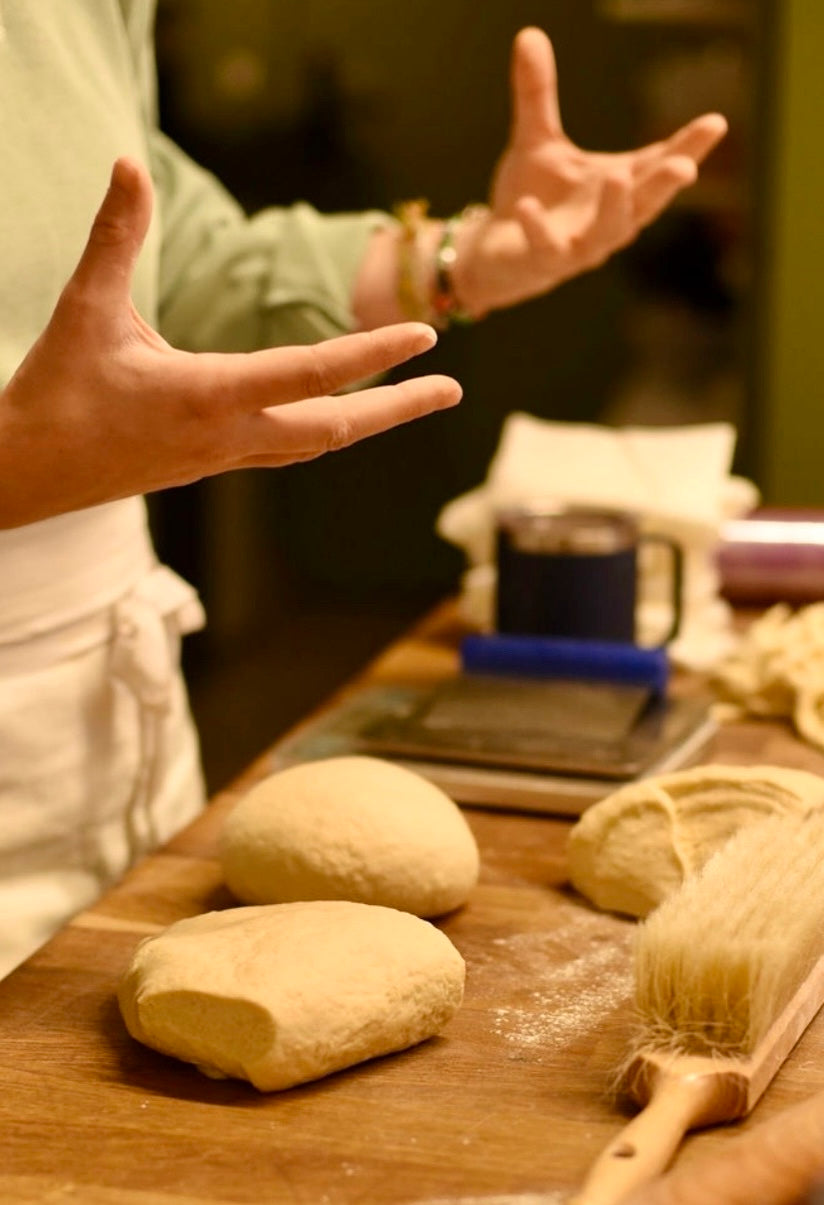 Pastry chef gesturing while explaining the lamination technique with blocks of croissant dough, a flour brush, and a scale on a wooden worktop during a croissant baking class at Maison Fleuret Paris