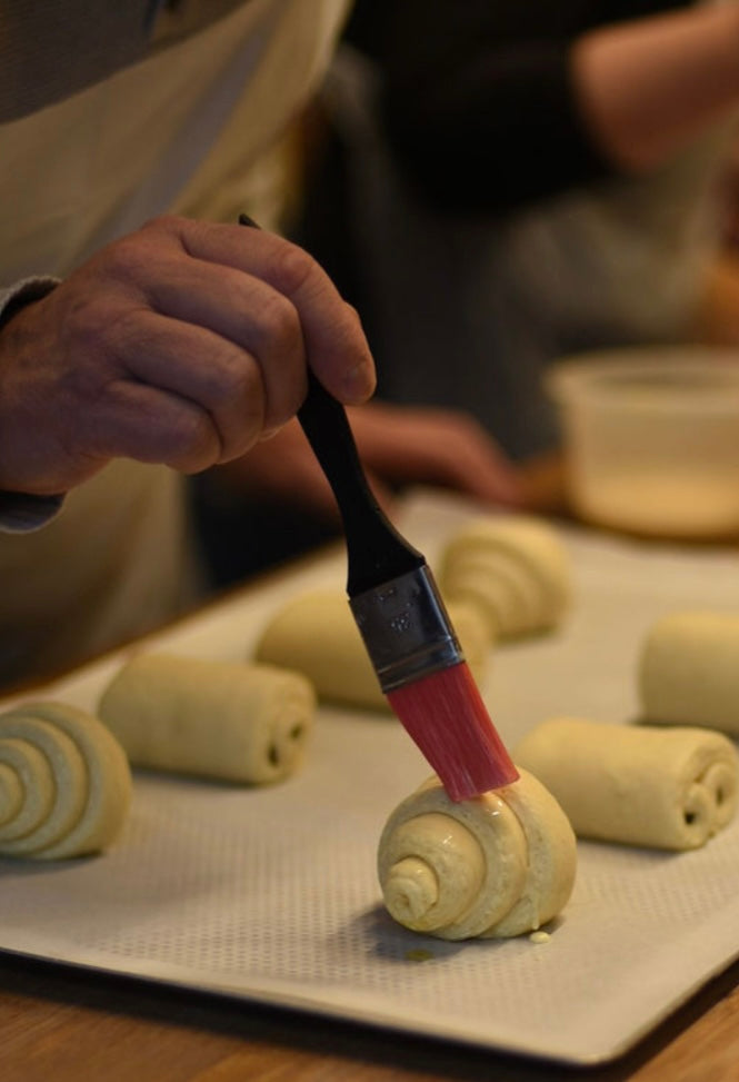Close-up of a hand brushing egg wash onto a shaped raw croissant with a silicone pastry brush before baking, with pains au chocolat and pains aux raisins on the tray during a croissant class at Maison Fleuret Paris