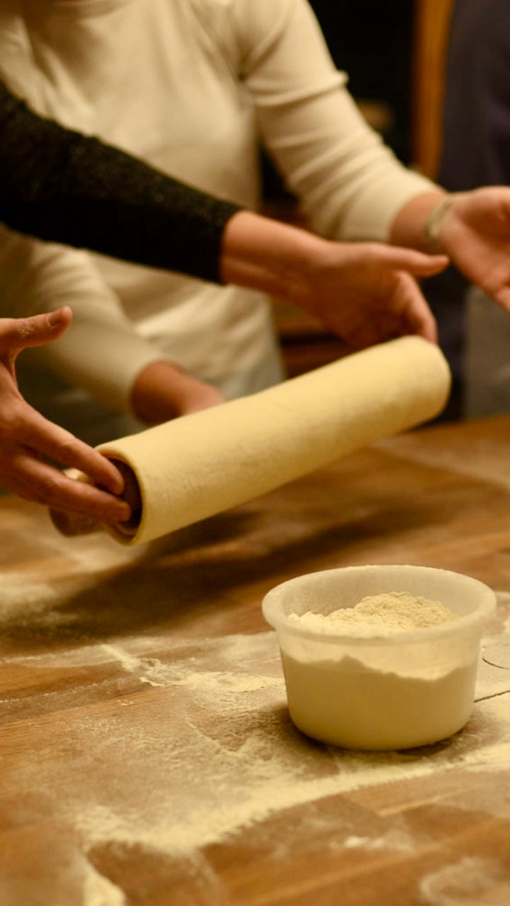 Participants handling a rolled sheet of laminated croissant dough with visible butter layers beside a bowl of flour on a wooden table during a hands-on croissant baking class at Maison Fleuret Paris
