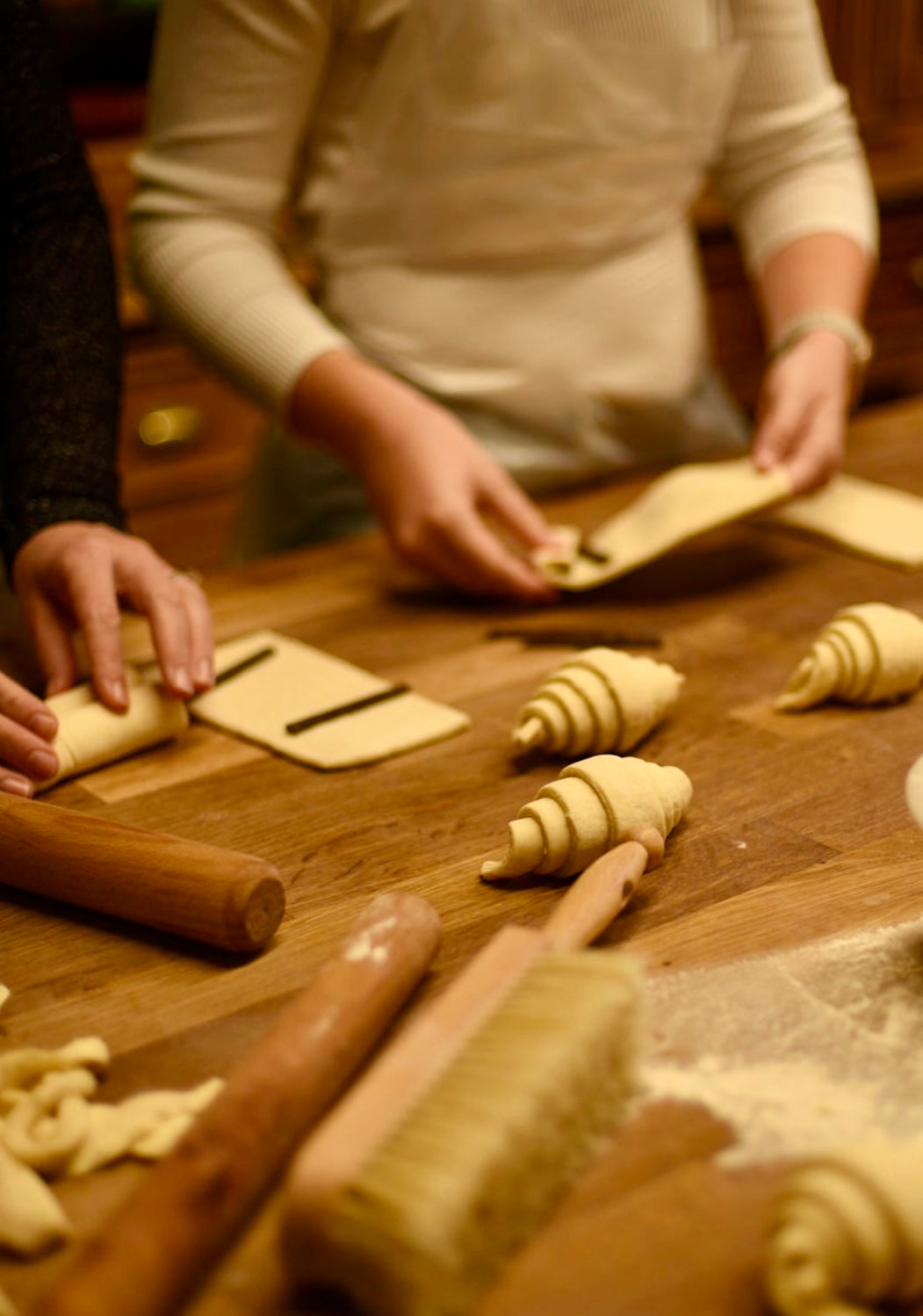 Participants rolling pains au chocolat with chocolate bars and shaping croissants on a wooden table surrounded by rolling pins and a flour brush during a hands-on croissant baking class at Maison Fleuret Paris