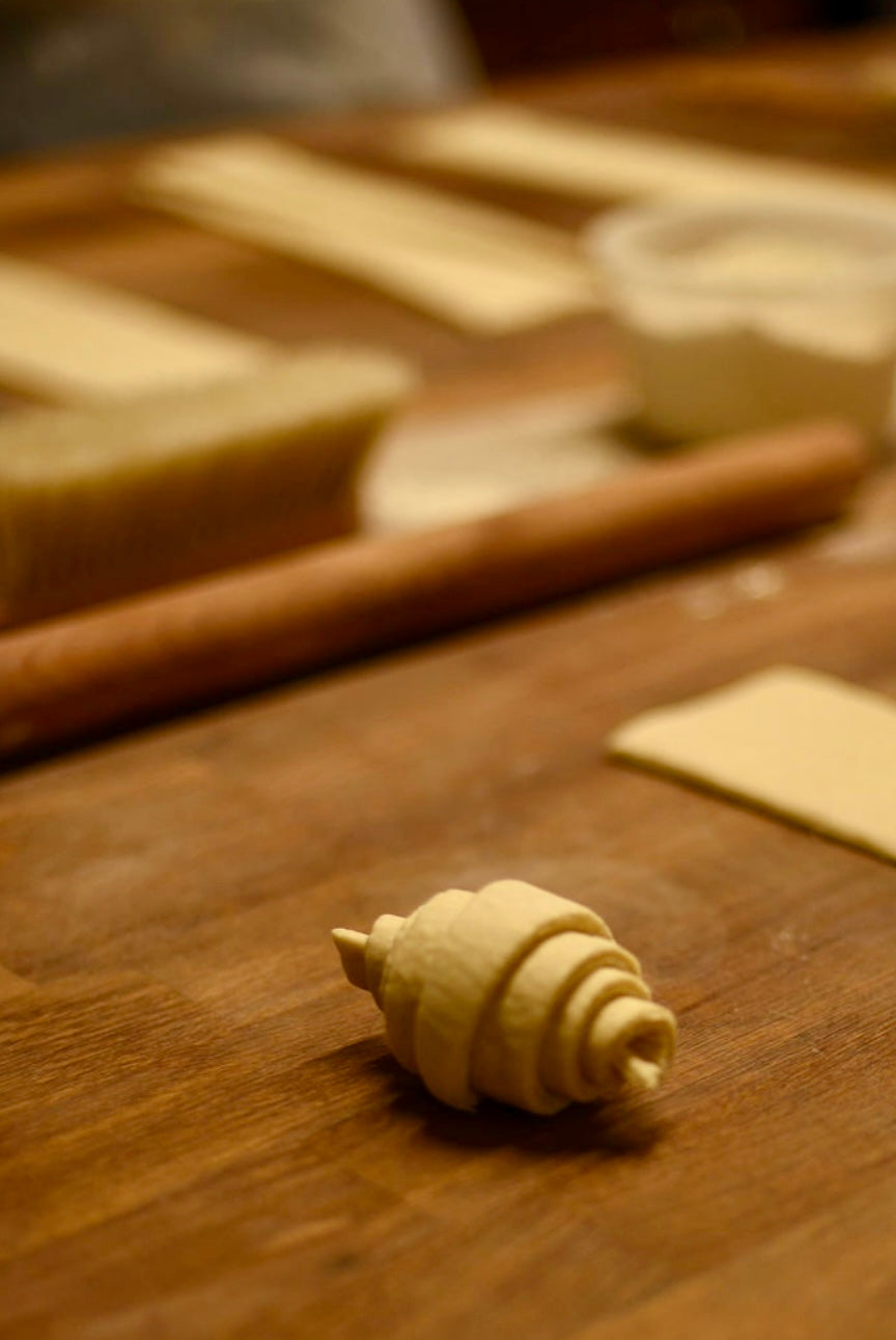 A perfectly shaped raw croissant dough with visible spiral layers resting on a wooden table before baking, with cut dough triangles and a baking tray in the background during a croissant class at Maison Fleuret Paris