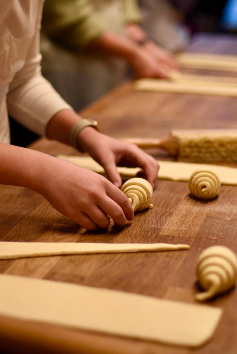 Participants rolling spiral-shaped pains aux raisins from laminated dough strips on a wooden table with cut dough triangles ready for shaping during a croissant baking class at Maison Fleuret Paris
