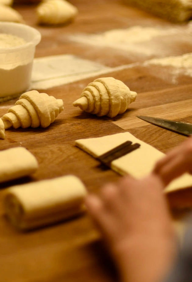 Shaped croissant dough and pain au chocolat being rolled with chocolate bars on a flour-dusted wooden table with a knife during a hands-on croissant baking class at Maison Fleuret Paris