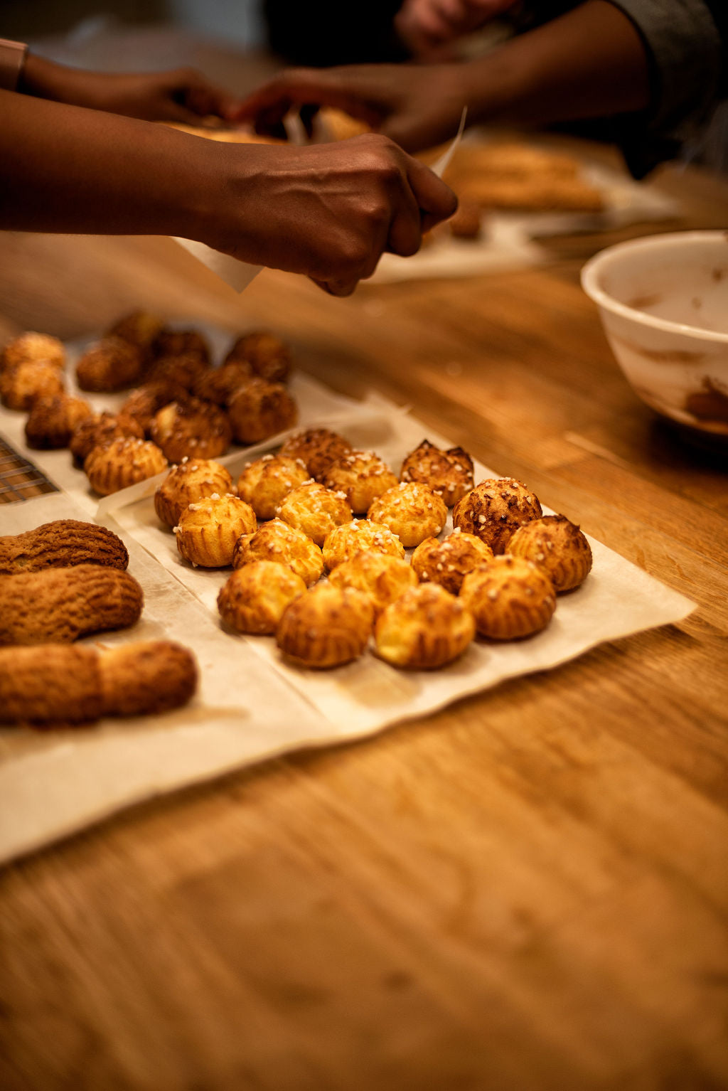 Freshly baked choux buns with pearl sugar and craquelin eclairs laid out on parchment paper with participants preparing fillings in the background during a Paris pastry workshop at Maison Fleuret