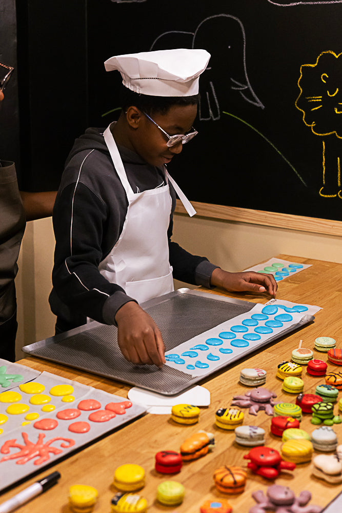 Young boy in chef hat and apron arranging blue macaron shells on a baking tray surrounded by colorful animal-shaped macarons, bees, octopus, ladybugs, and rainbow shells during a kids macaron class at Maison Fleuret Paris