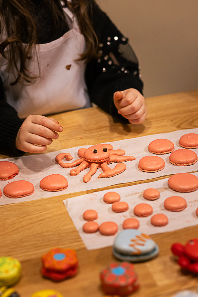 Child in apron admiring a creative octopus-shaped macaron with chocolate eyes surrounded by orange macaron shells and colorful decorated cookies during a kids macaron class at Maison Fleuret Paris