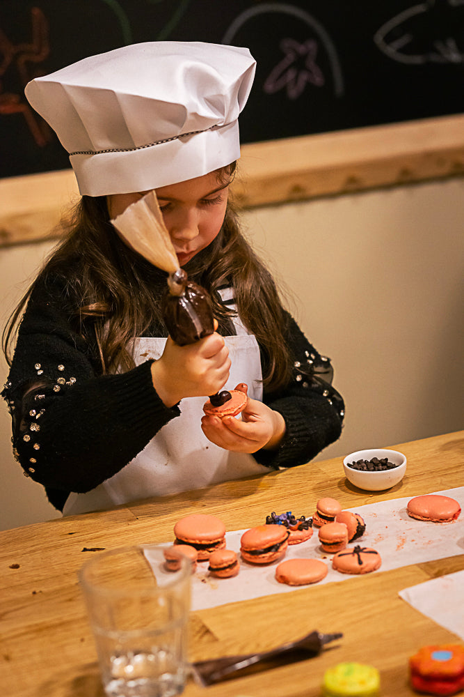 Young girl wearing a white chef hat and apron piping chocolate filling onto orange macaron shells with a pastry bag at a wooden table during a kids macaron baking class at Maison Fleuret Paris