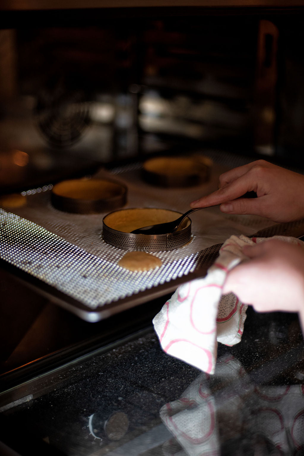Hand checking shortcrust tart shells blind-baking in perforated rings on a perforated baking tray inside the oven during a lemon meringue tart class at Maison Fleuret Paris
