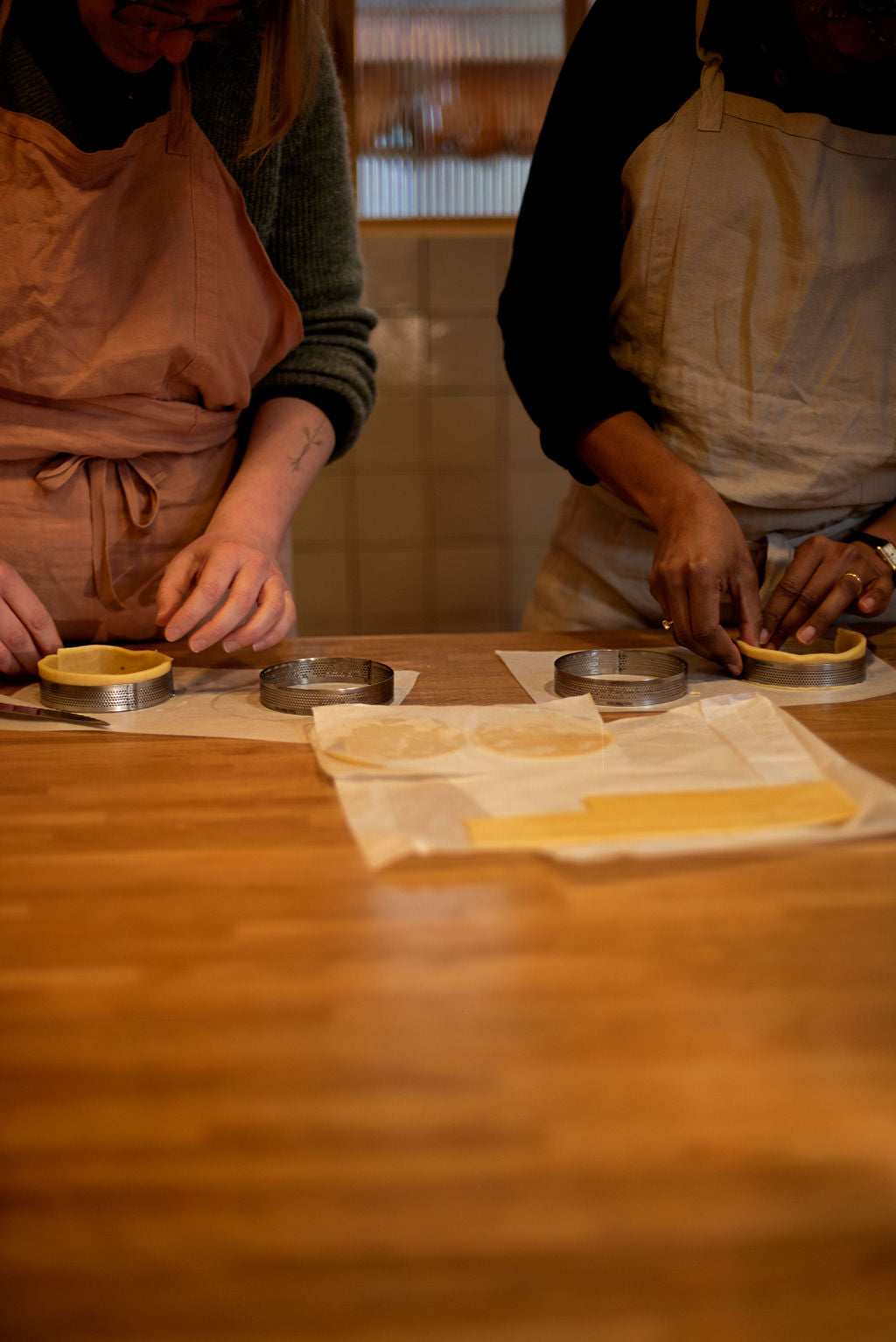 Two participants in aprons lining perforated tart rings with shortcrust pastry dough on a wooden table during a small group lemon meringue tart class at Maison Fleuret Paris