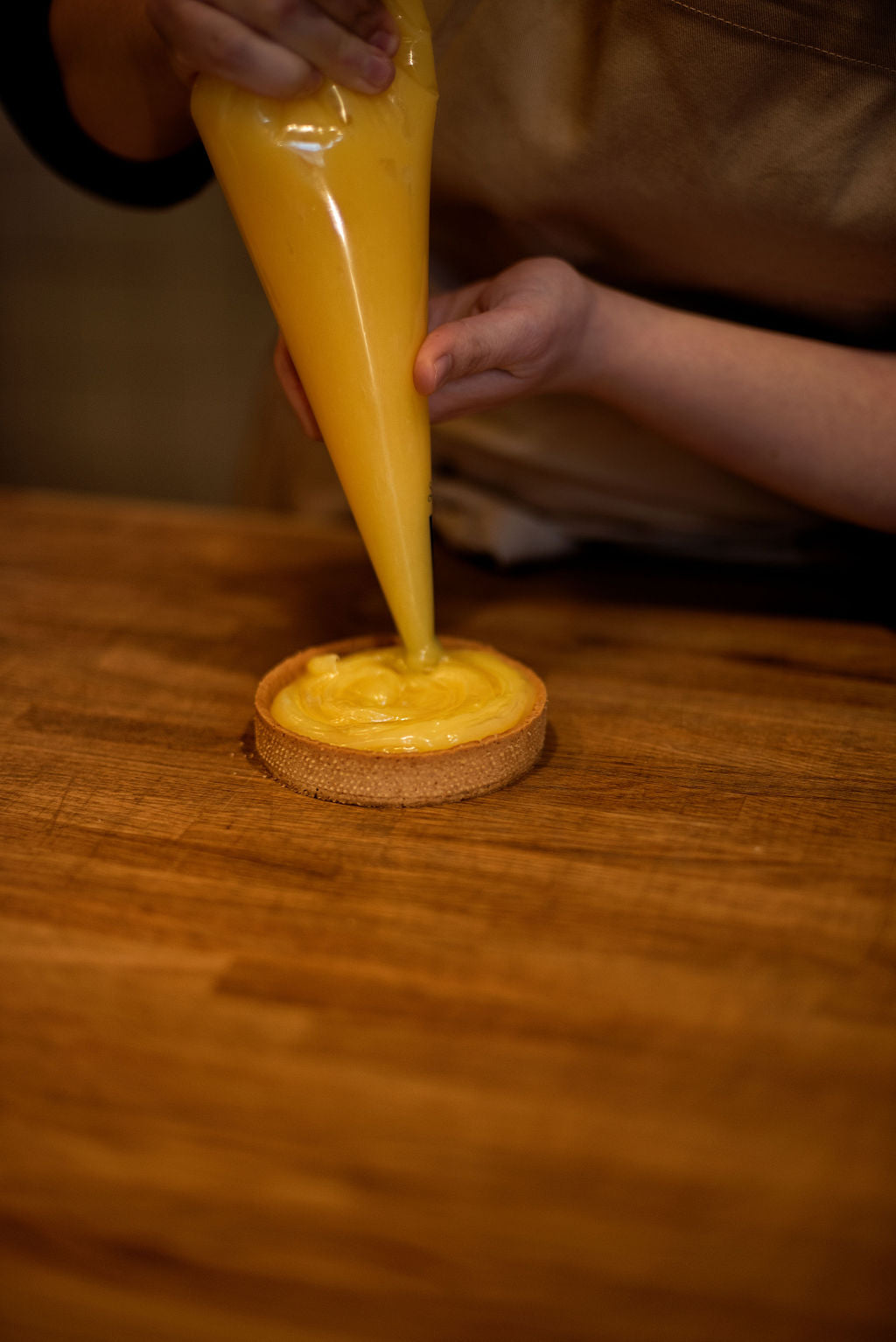 Piping vibrant lemon curd from a pastry bag into a baked tart shell on a wooden table during a hands-on lemon meringue tart class at Maison Fleuret Paris