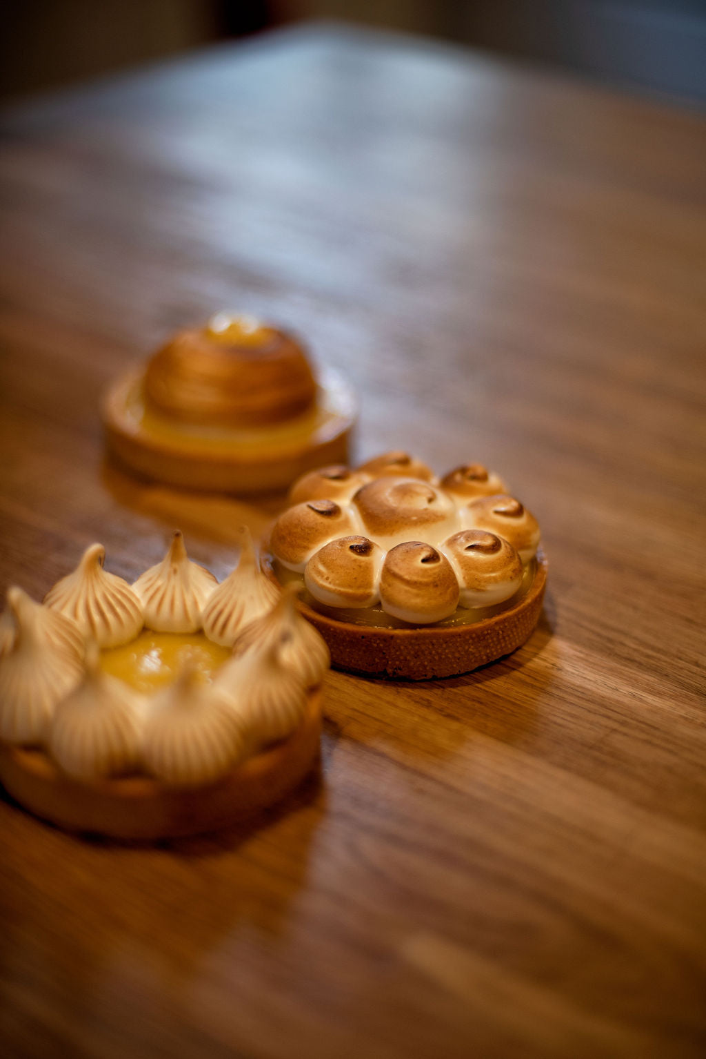 Three French lemon meringue tarts with different meringue styles, piped peaks, torched rosettes, and a smooth dome, displayed on a wooden table during a pastry class at Maison Fleuret Paris