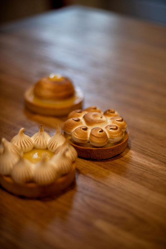 Three French lemon meringue tarts with different meringue styles, piped peaks, torched rosettes, and a smooth dome, displayed on a wooden table during a pastry class at Maison Fleuret Paris