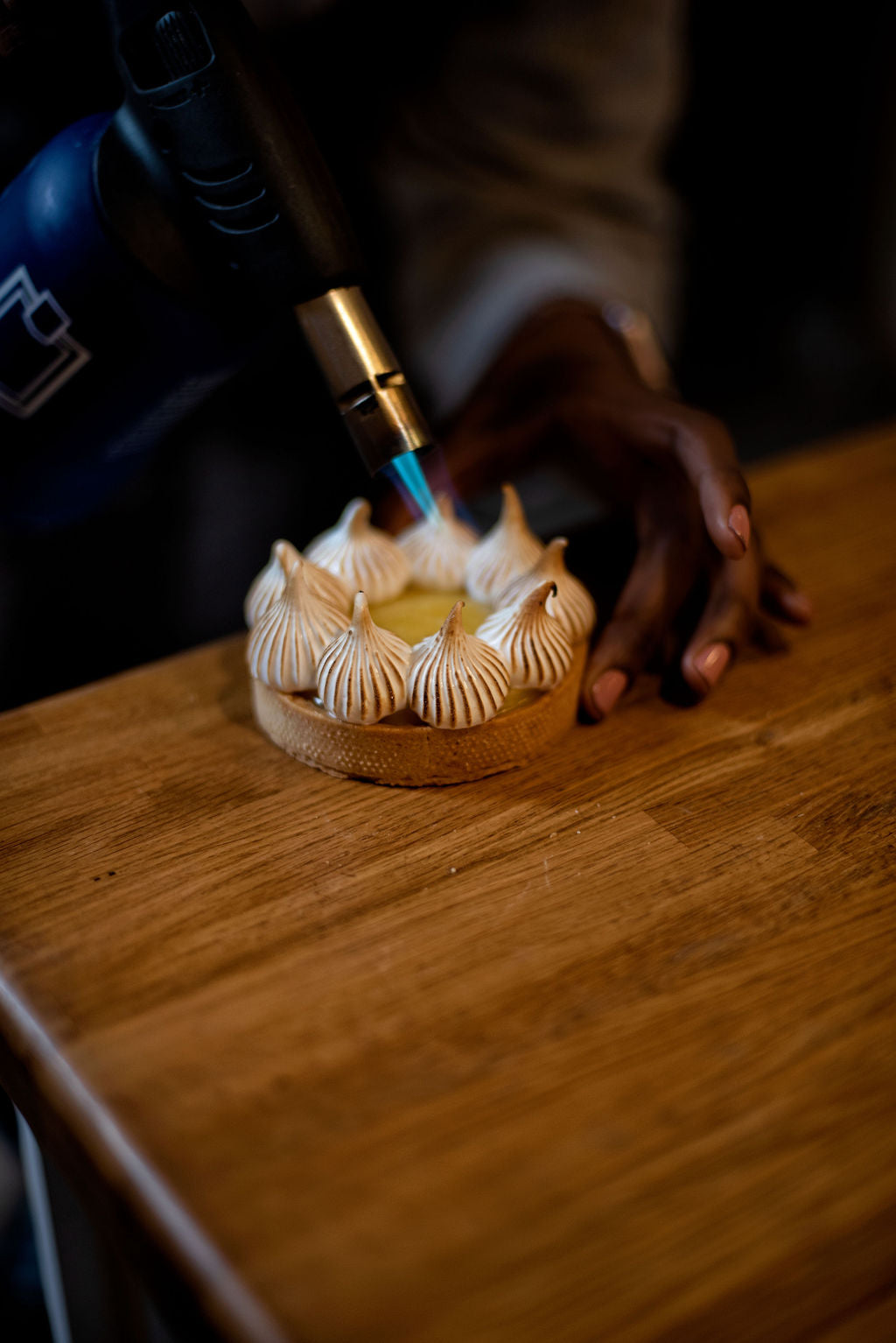 Participant torching elegant piped meringue peaks on a lemon tart with a kitchen blowtorch on a wooden table during a hands-on pastry class at Maison Fleuret Paris