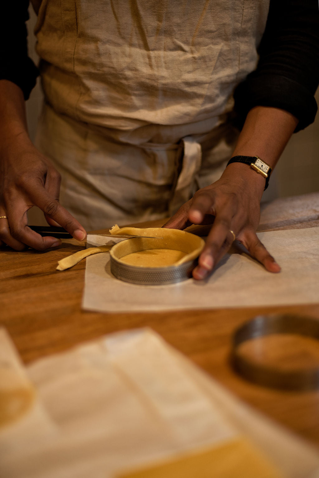 Participant in apron trimming excess shortcrust pastry from a perforated tart ring with a knife to create a clean tart shell during a lemon meringue class at Maison Fleuret Paris