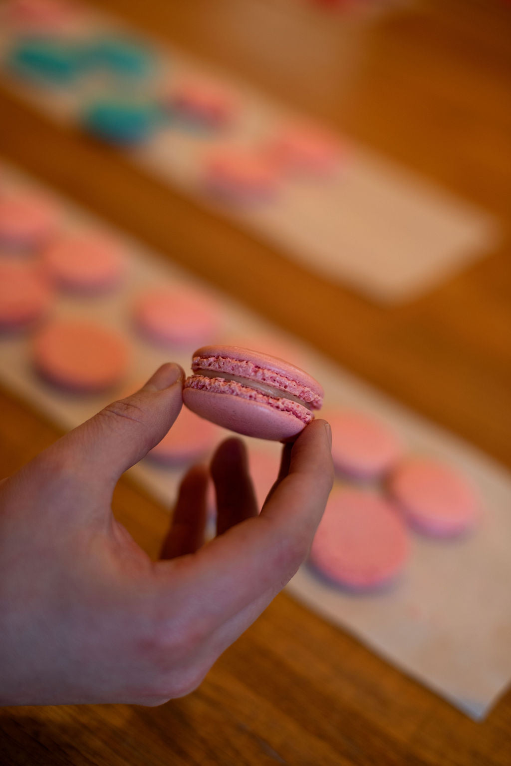 Hand holding a freshly made pink macaron during a baking class at Maison Fleuret Paris, with rows of colorful macarons on a wooden table