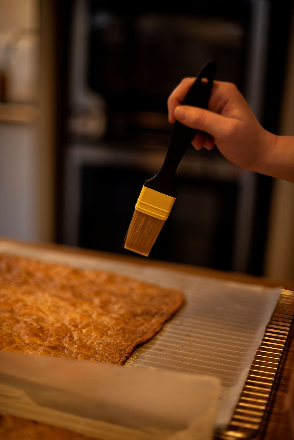 Hand brushing a golden caramelized puff pastry sheet on a cooling rack with a pastry brush in front of the oven during a Napoleon millefeuille class at Maison Fleuret Paris