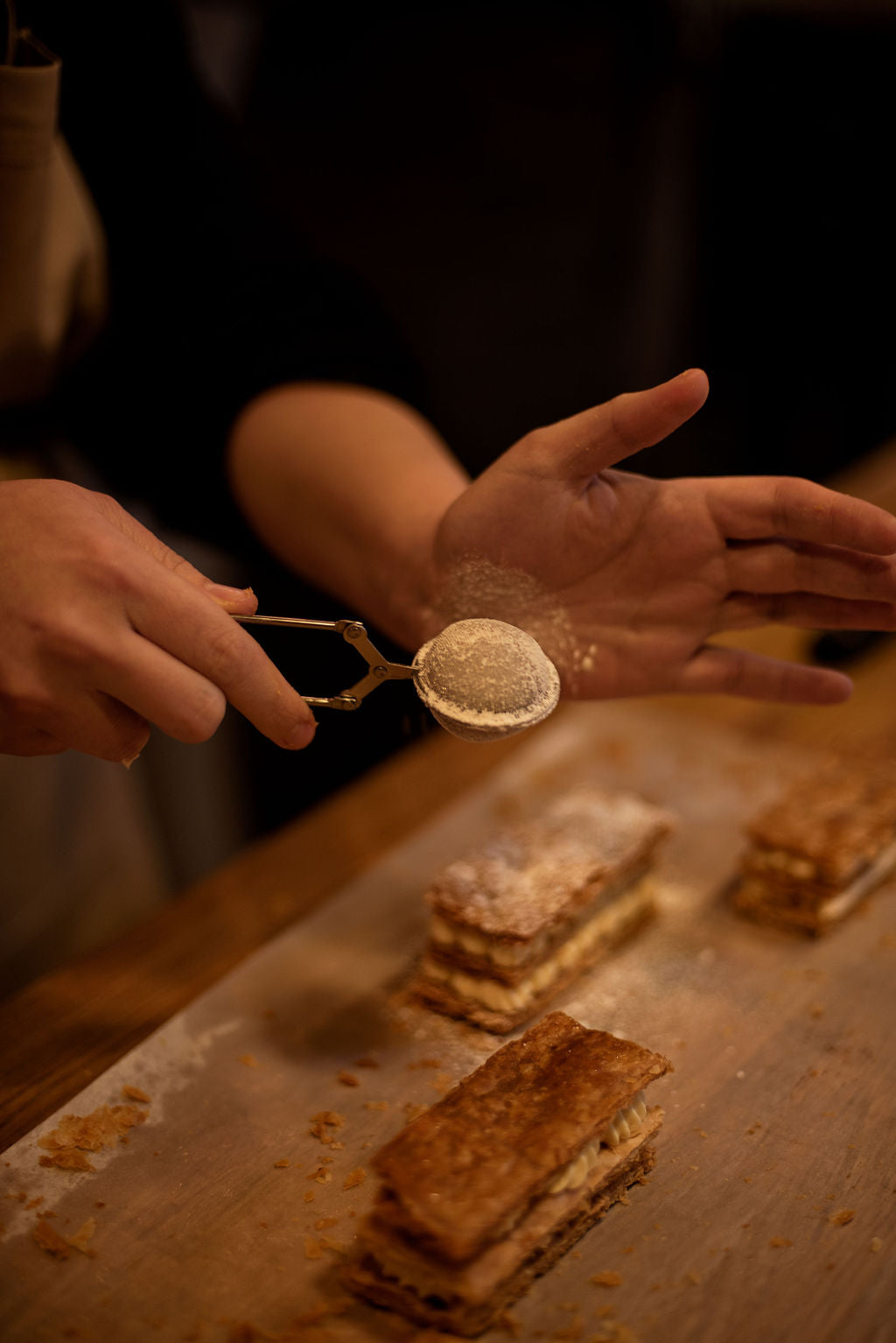 Hands dusting powdered sugar through a small sieve over finished millefeuille Napoleons as a final decoration during a pastry class at Maison Fleuret Paris