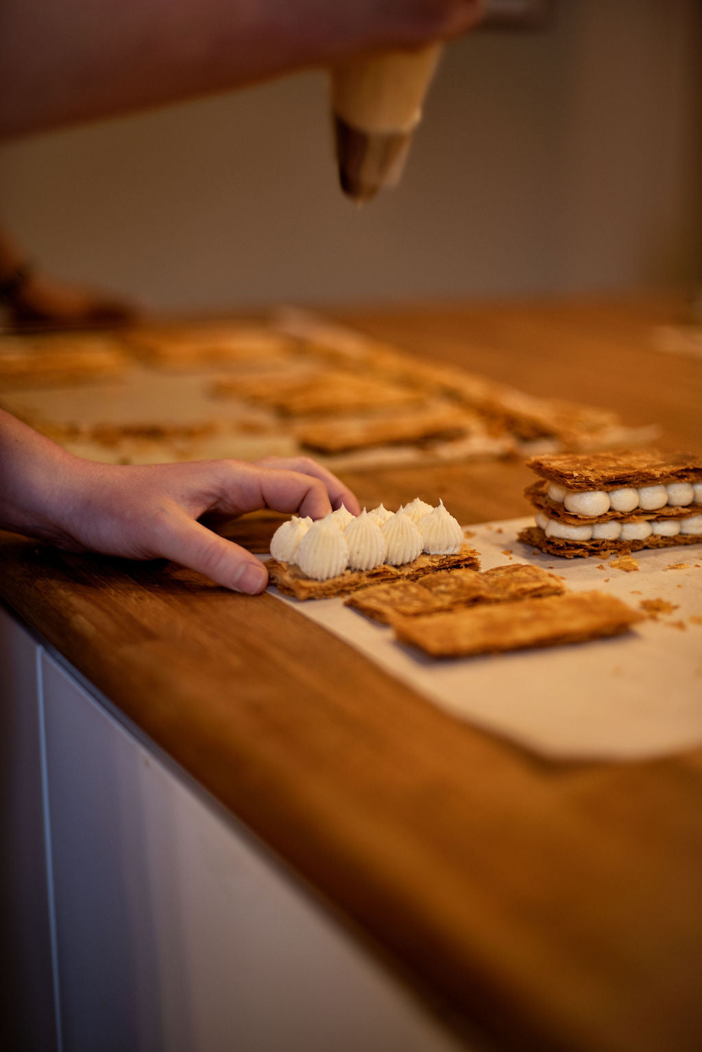 Hand positioning a layer of caramelized puff pastry next to piped pastry cream rosettes with assembled millefeuilles in the background during a Napoleon class at Maison Fleuret Paris