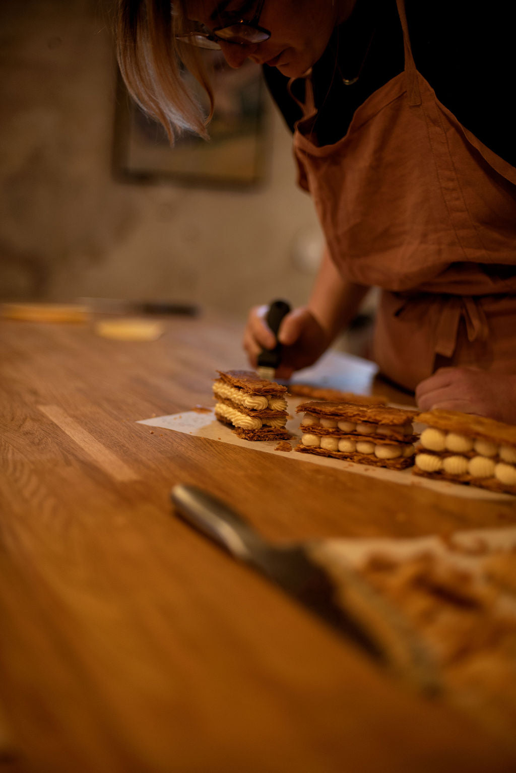 Participant in apron torching the top of assembled millefeuilles with a kitchen blowtorch to caramelize the puff pastry layers during a Napoleon class at Maison Fleuret Paris