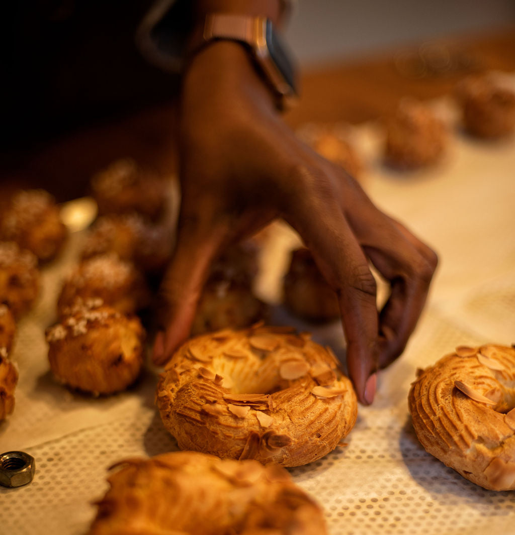 Hand reaching for freshly baked golden Paris-Brest choux rings topped with slivered almonds on a perforated baking mat alongside choux buns during a pastry class at Maison Fleuret Paris