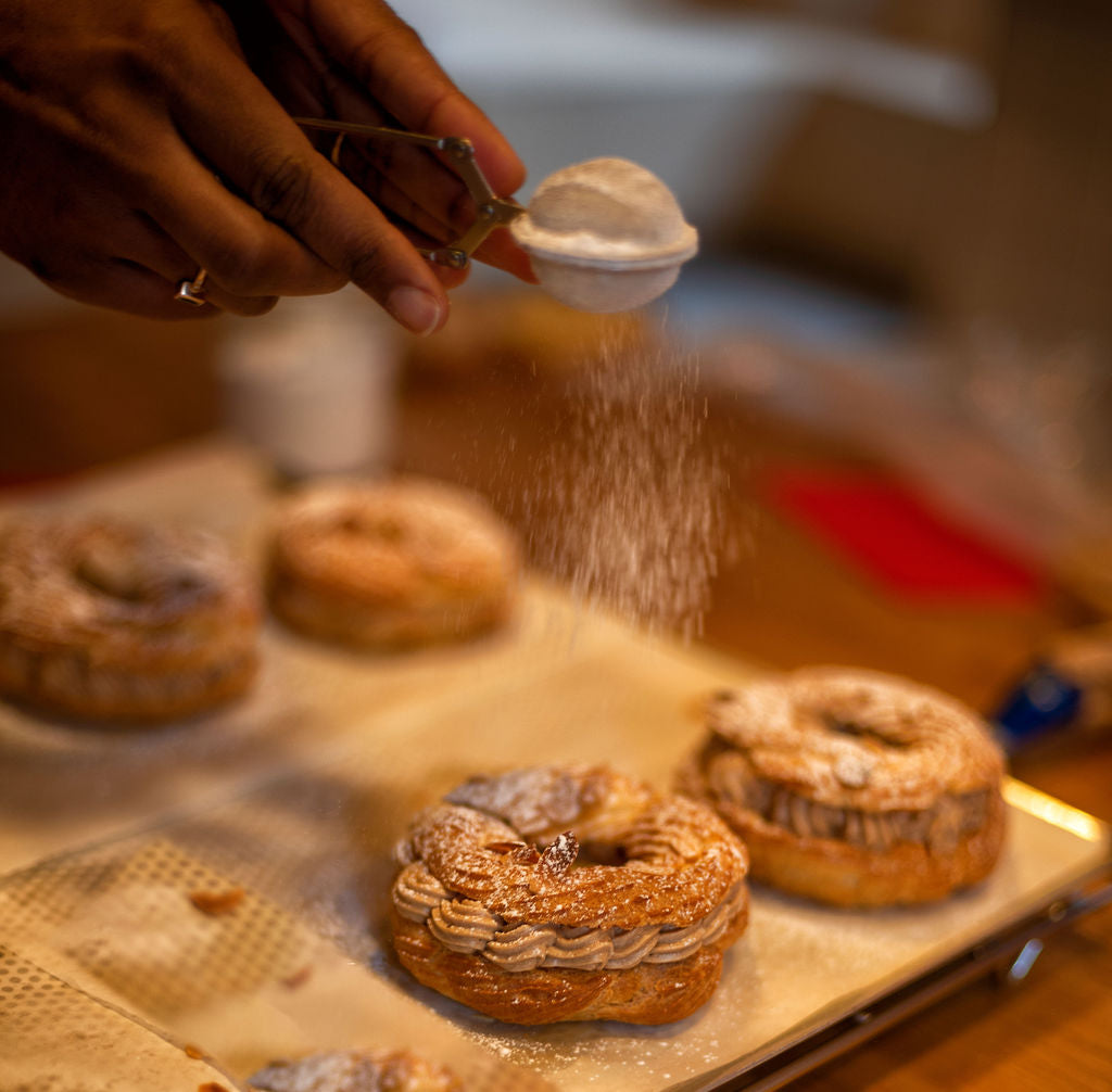 Hand dusting powdered sugar through a small sieve over finished Paris-Brest pastries filled with piped praline mousseline cream on a baking tray during a pastry class at Maison Fleuret Paris