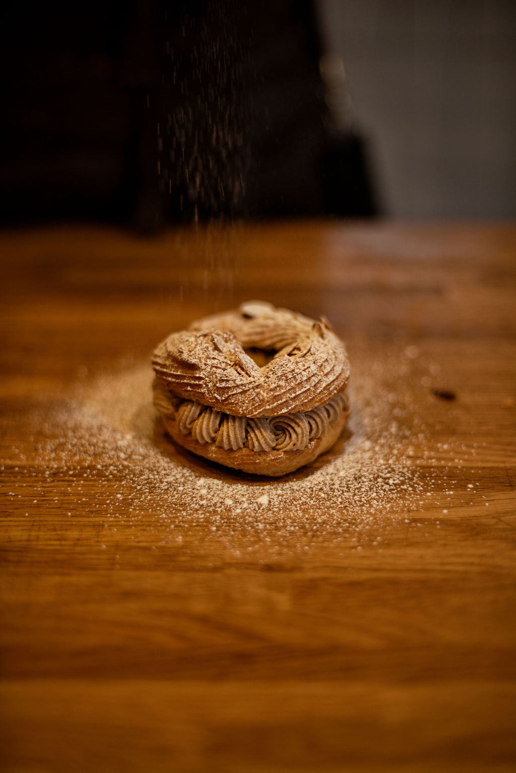 Finished Paris-Brest pastry filled with praline mousseline cream being showered with powdered sugar falling from above on a wooden table — made during a pastry class at Maison Fleuret Paris