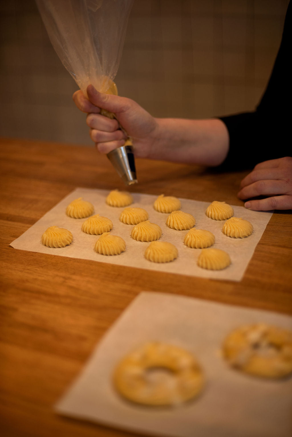 Participant piping rows of ridged choux buns with a star-tipped pastry bag onto parchment paper alongside a Paris-Brest ring during a hands-on pastry class at Maison Fleuret Paris