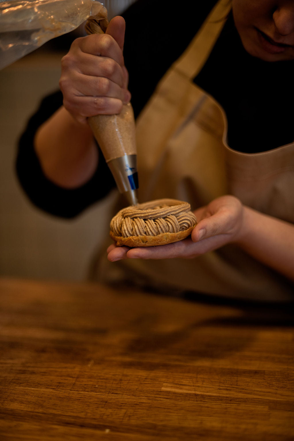 Participant in apron piping hazelnut praline mousseline cream in a spiral pattern into a choux ring held in hand during a Paris-Brest pastry class at Maison Fleuret Paris