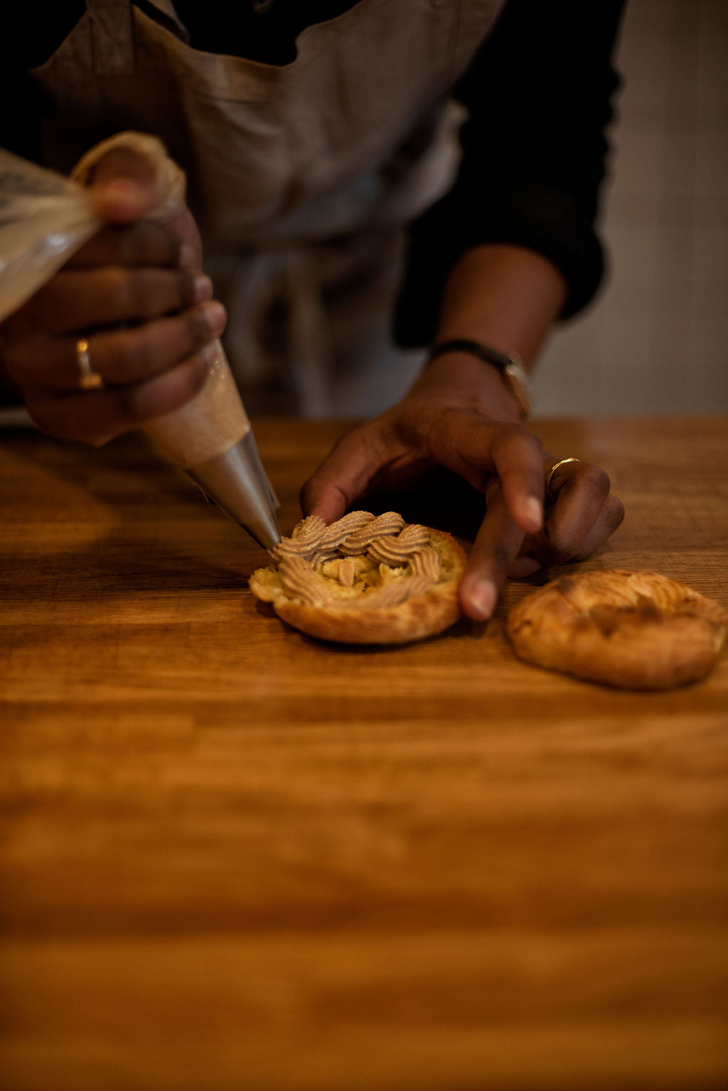 Participant piping hazelnut praline mousseline cream in a decorative wave pattern into a sliced Paris-Brest choux ring on a wooden table during a pastry class at Maison Fleuret Paris
