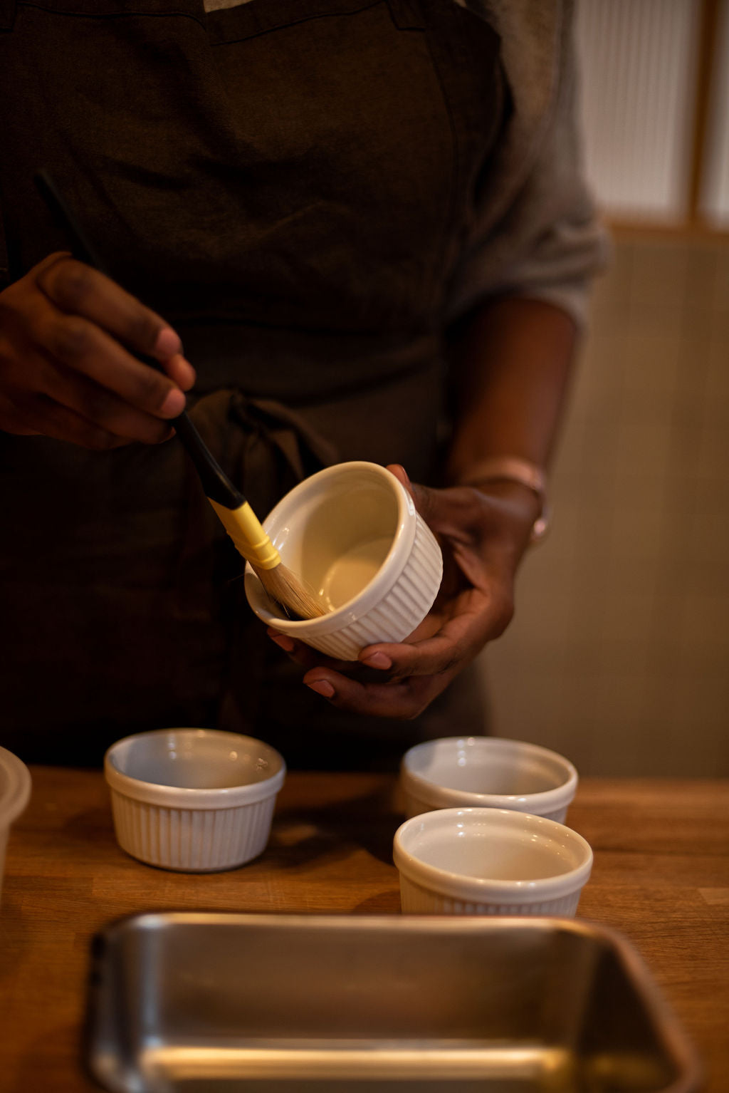 Participant in apron brushing butter inside a white ceramic ramekin with a pastry brush, the key preparation step for a perfect souffle rise, during a class at Maison Fleuret Paris near Notre-Dame