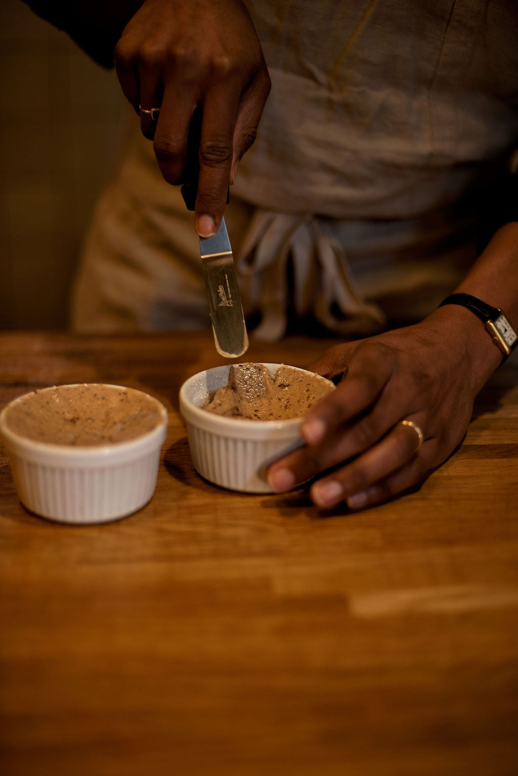 Participant leveling a risen chocolate souffle in a white ramekin with a palette knife on a wooden table during a French souffle baking class at Maison Fleuret Paris near Notre-Dame