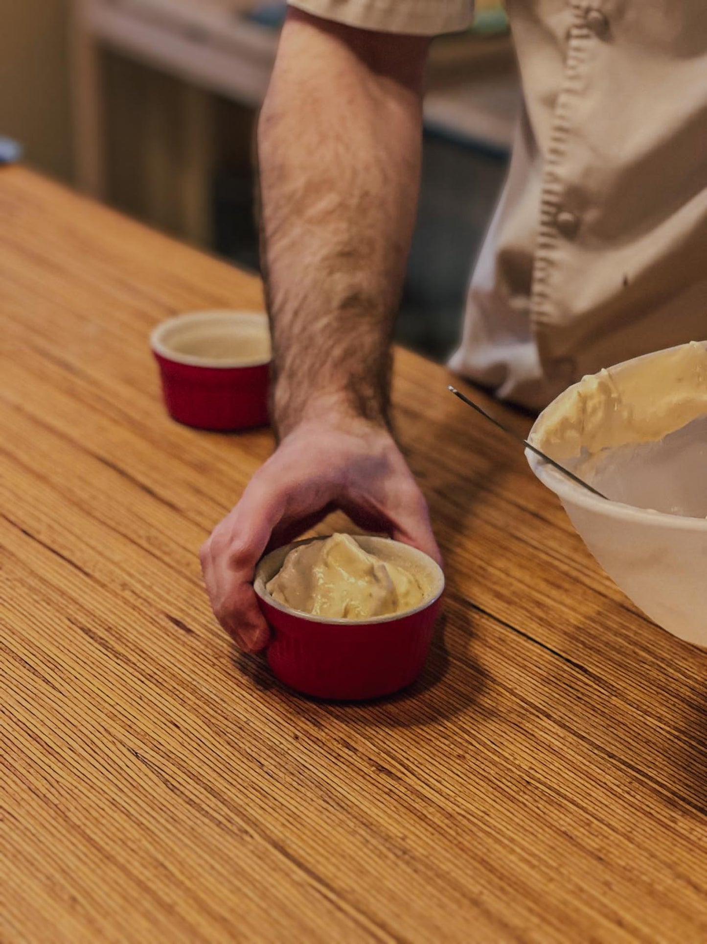 Participant filling a red ceramic ramekin with souffle batter from a mixing bowl on a wooden worktop during a hands-on French souffle baking class at Maison Fleuret Paris near Notre-Dame
