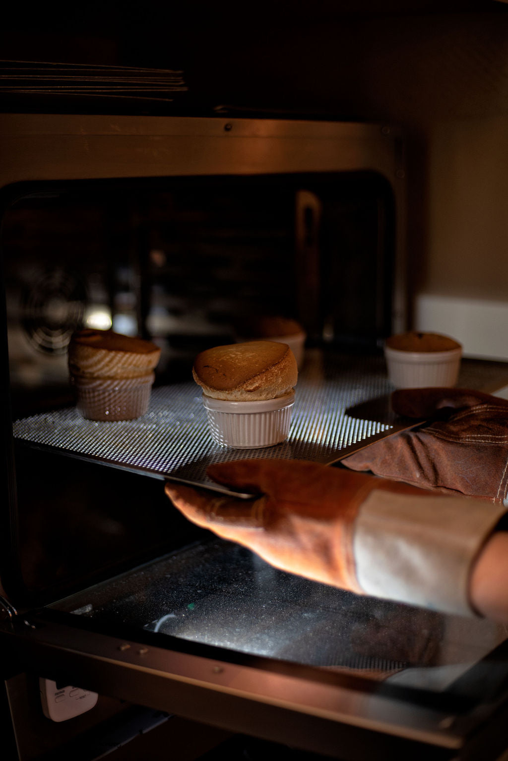 Hand with oven mitt pulling a tray of perfectly risen souffles in white ramekins out of the oven during a French souffle baking class at Maison Fleuret Paris near Notre-Dame