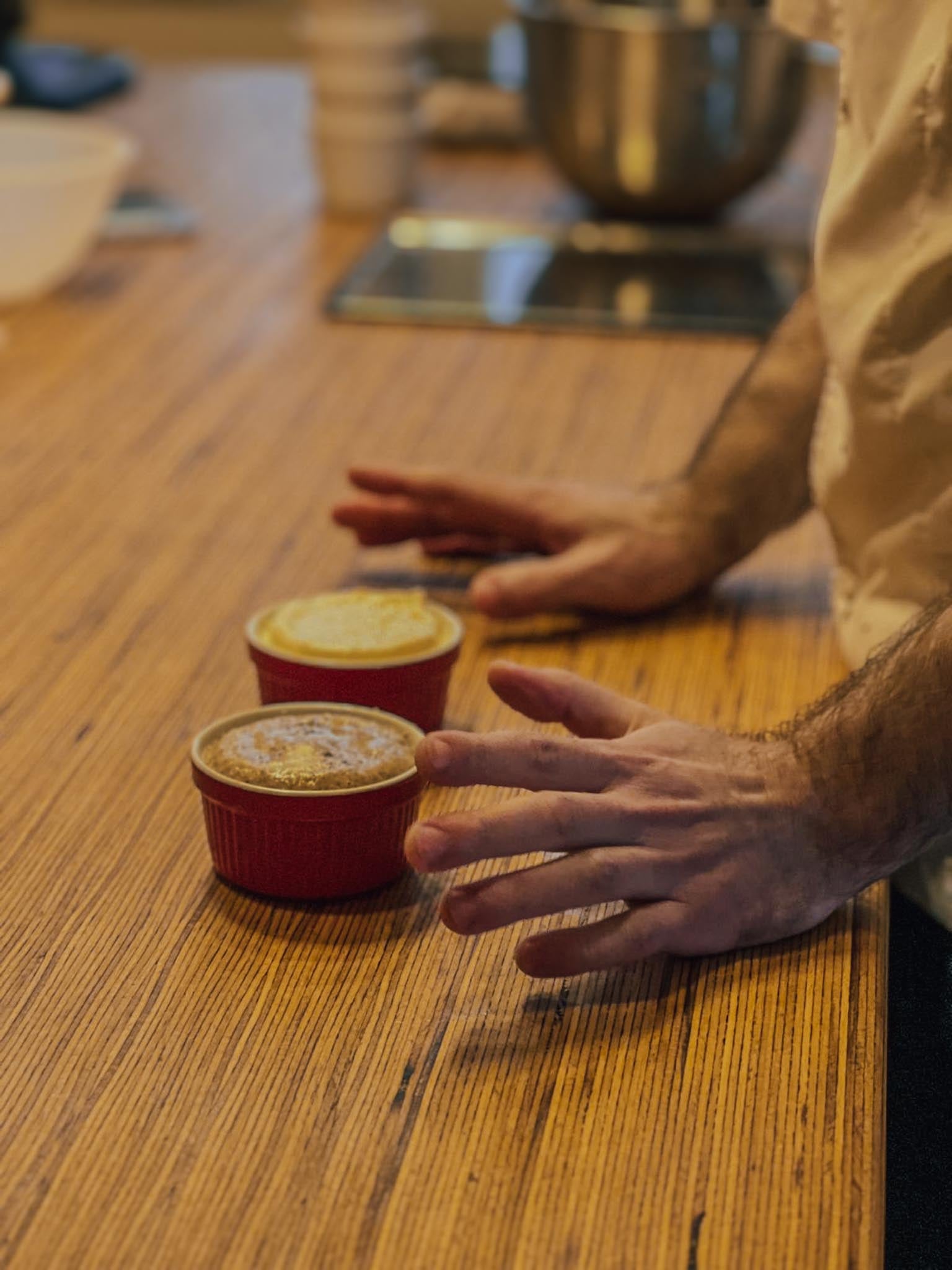 Two freshly baked souffles in red ceramic ramekins dusted with powdered sugar on a wooden worktop with the pastry chef's hand nearby during a souffle class at Maison Fleuret Paris near Notre-Dame