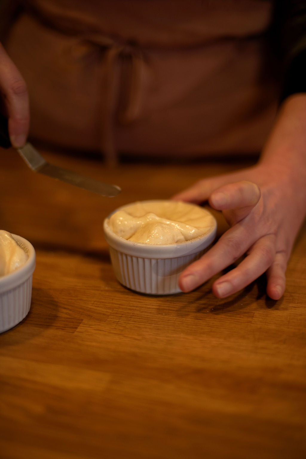 Participant in apron placing a perfectly risen golden souffle in a white ramekin on a wooden table with a palette knife during a French souffle baking class at Maison Fleuret Paris near Notre-Dame